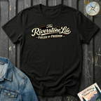 A black short-sleeve graphic T-shirt laid flat on a dark textured surface, with a vintage-style cream and tan slogan reading 'The Riverstone Life FUELED BY FREEDOM.' A denim jacket corner, a compass illustration, and a Rocky Mountain National Park stamp-style badge are placed around it as styling props.