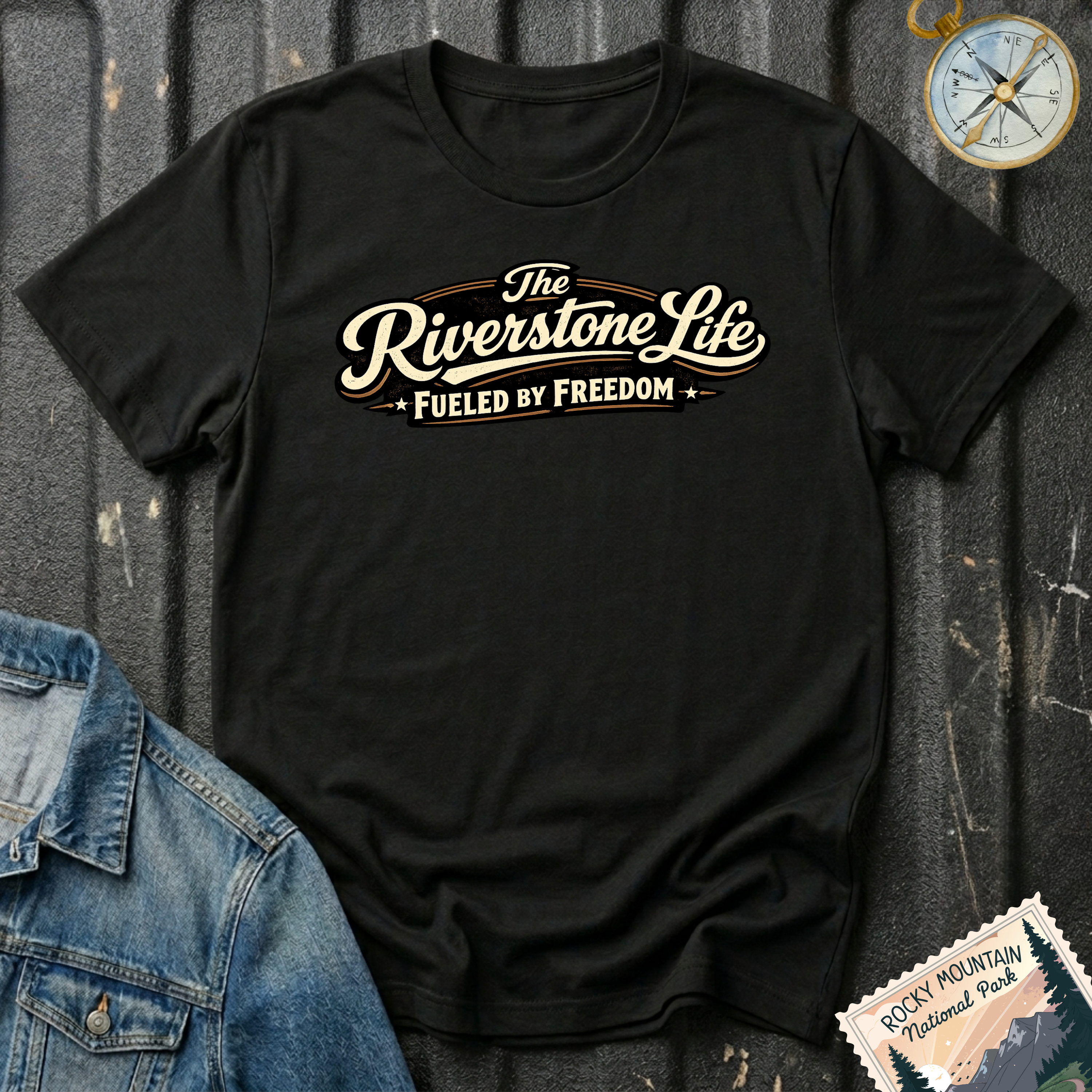A black short-sleeve graphic T-shirt laid flat on a dark textured surface, with a vintage-style cream and tan slogan reading 'The Riverstone Life FUELED BY FREEDOM.' A denim jacket corner, a compass illustration, and a Rocky Mountain National Park stamp-style badge are placed around it as styling props.