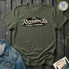 A military green short-sleeve t-shirt with a retro-style graphic reading 'The Riverstone Life FUELED BY FREEDOM' in cream and brown lettering, styled in a flat-lay arrangement with a denim jacket, a compass, and a Rocky Mountain National Park stamp on a dark textured background.