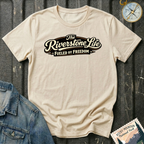 A natural graphic t-shirt laid flat on a dark tailgate background, styled with a denim jacket corner, a compass, and a Rocky Mountain National Park postage stamp. The t-shirt features a retro-style graphic that reads 'The Riverstone Life FUELED BY FREEDOM'.