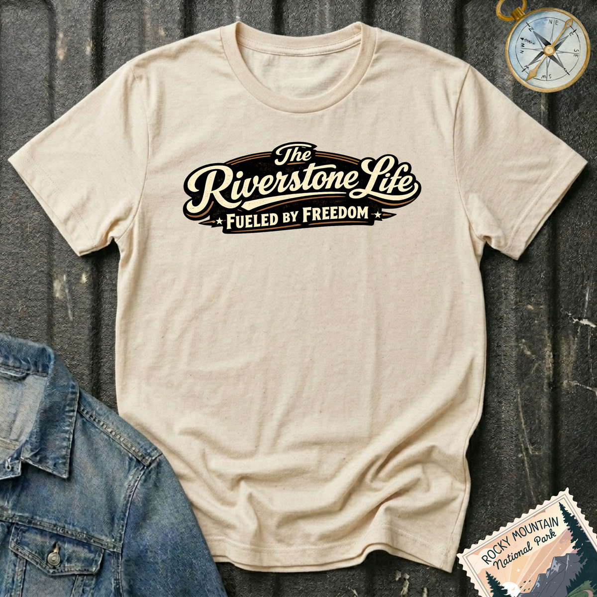 A natural graphic t-shirt laid flat on a dark tailgate background, styled with a denim jacket corner, a compass, and a Rocky Mountain National Park postage stamp. The t-shirt features a retro-style graphic that reads 'The Riverstone Life FUELED BY FREEDOM'.