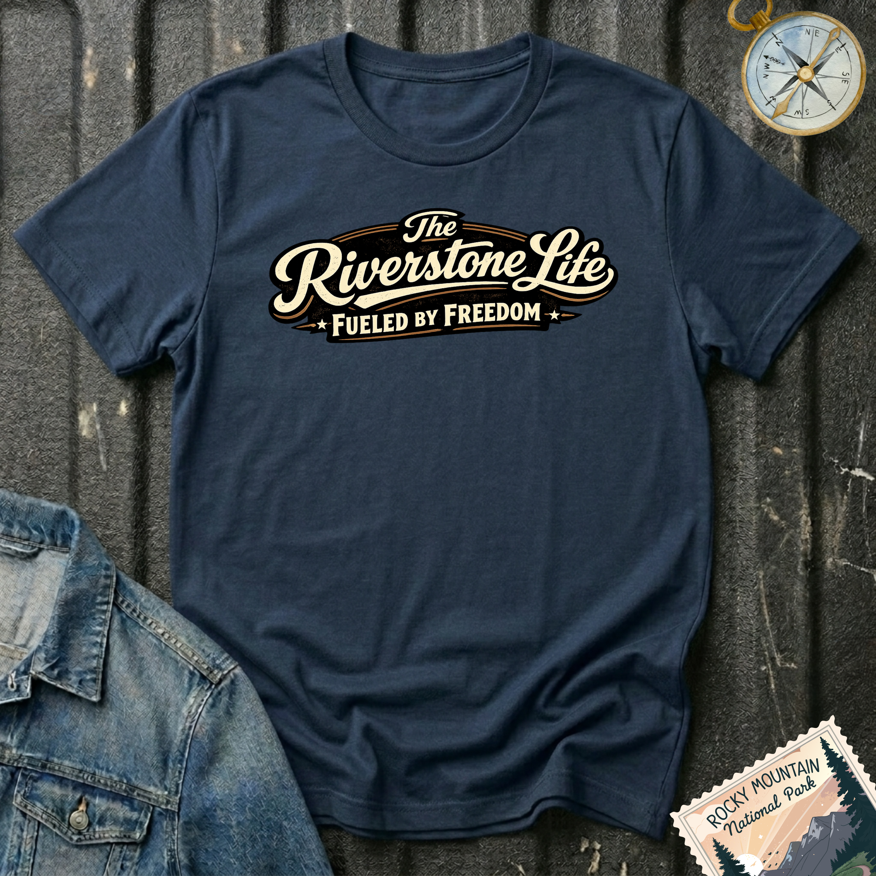 A navy graphic t-shirt laid flat on a dark textured background, styled with a denim jacket corner, a compass, and a Rocky Mountain National Park stamp. The t-shirt features a retro-style cream and brown graphic that reads 'The Riverstone Life FUELED BY FREEDOM'.