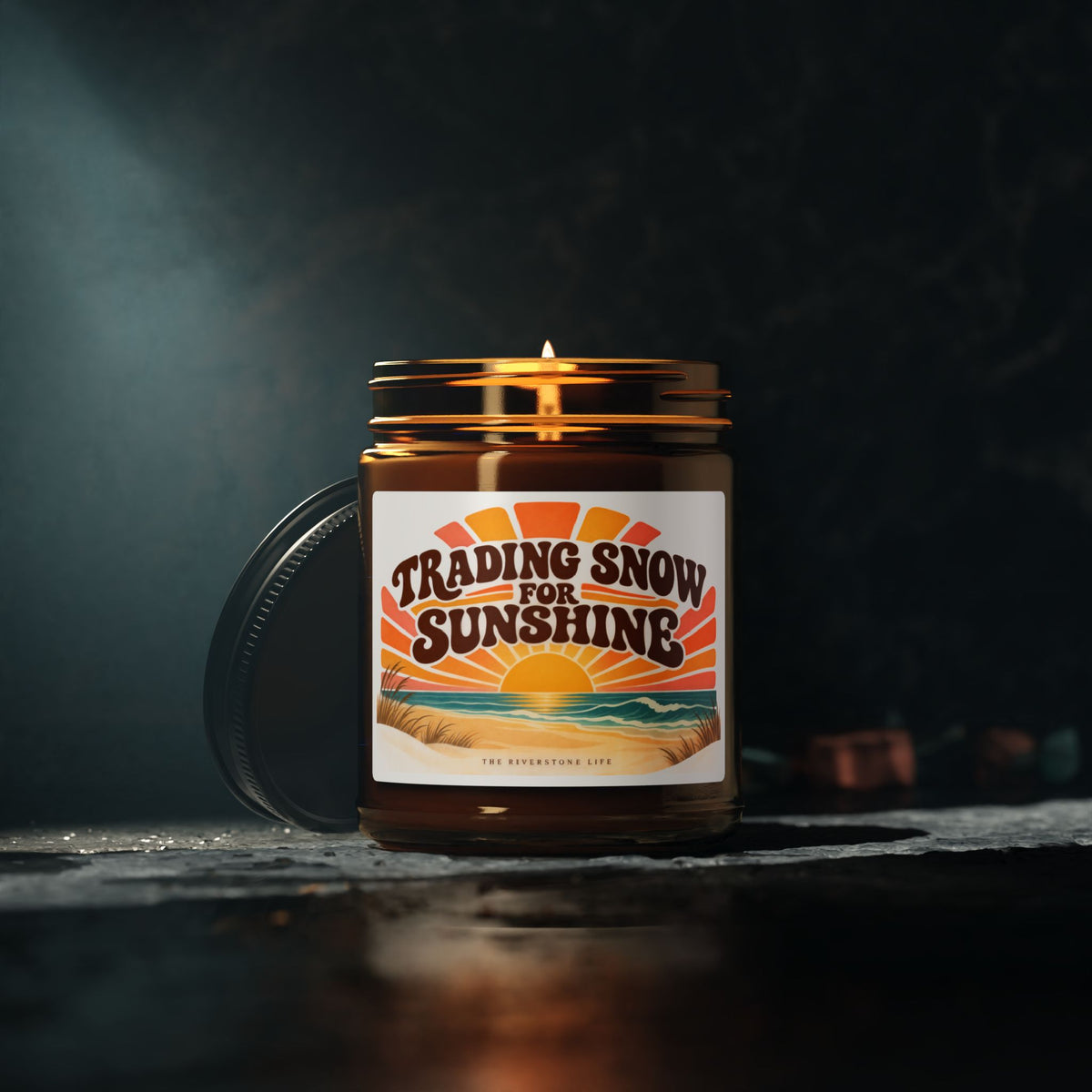 An amber glass jar candle with a lit wick and a black lid resting beside it on a dark surface. The front label reads “Trading Snow for Sunshine” with a sunset beach illustration and small text at the bottom that appears to say “The Riverstone Life.