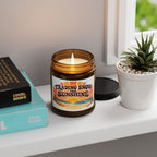 Candle labeled 'Trading Snow for Sunshine' with a small The Riverstone Life name at the bottom on a shelf with books and a plant