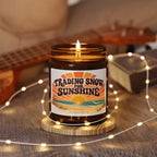 A lit amber glass jar candle sits on a wooden coaster in a cozy indoor setting, surrounded by warm fairy lights. The candle features a retro-style label that reads 'TRADING SNOW FOR SUNSHINE' with an orange and yellow sunset over a beach with a small 'The Riverstone Life' name on the bottom