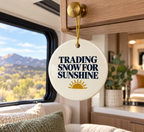 A round ceramic ornament hanging from a wooden cabinet inside an RV, featuring the text 'TRADING SNOW FOR SUNSHINE' in navy blue with a gold sun graphic below, set against a scenic mountain background visible through the window.
