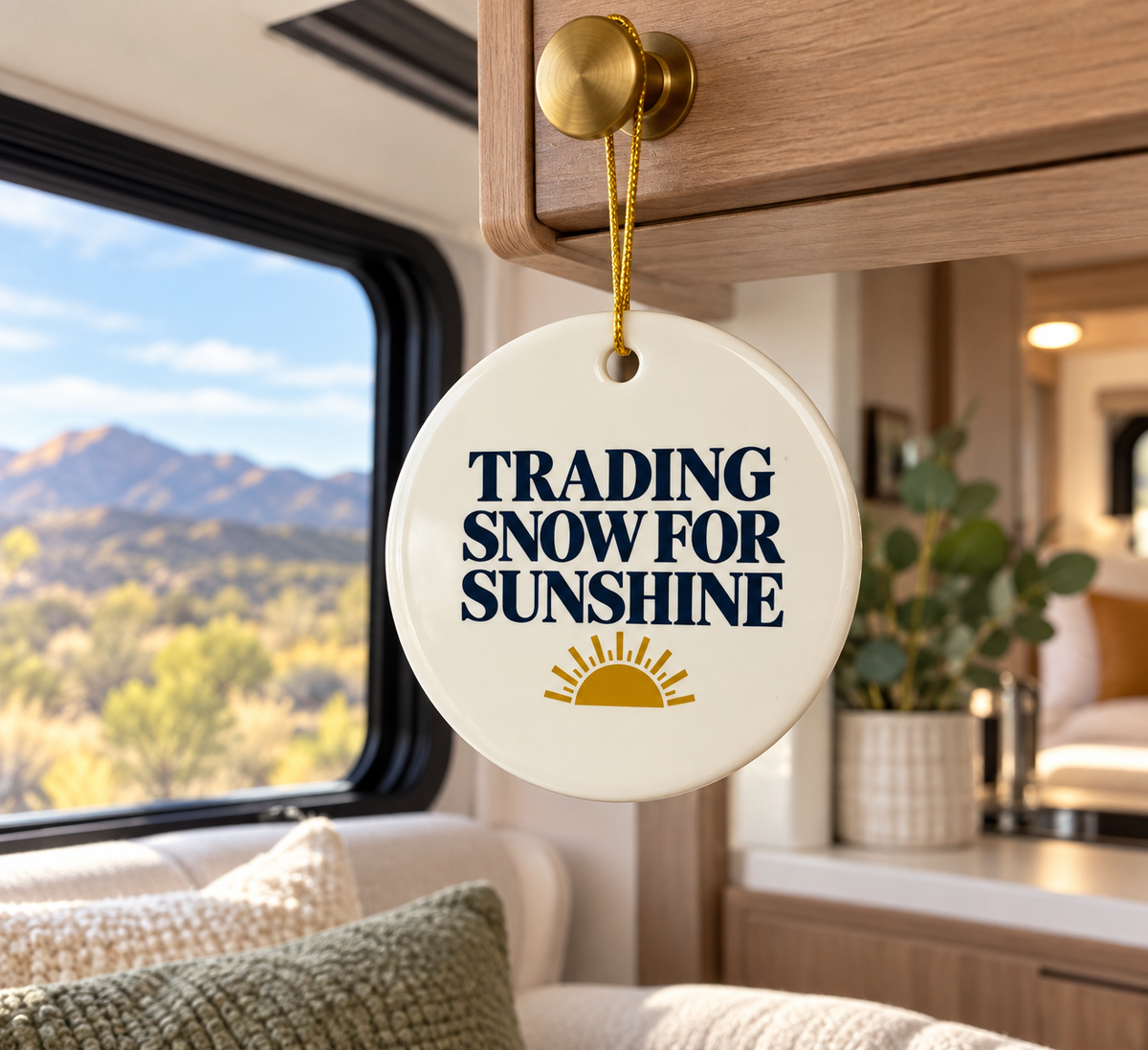 A round ceramic ornament hanging from a wooden cabinet inside an RV, featuring the text 'TRADING SNOW FOR SUNSHINE' in navy blue with a gold sun graphic below, set against a scenic mountain background visible through the window.