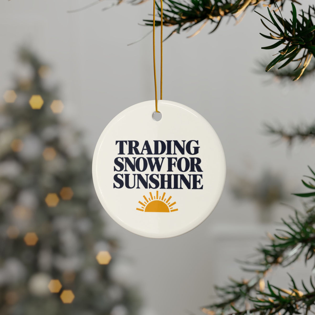 A round white ceramic Christmas ornament hanging from a gold string on a Christmas tree branch. The ornament features the text 'TRADING SNOW FOR SUNSHINE' in dark navy blue letters above a small yellow sun graphic, with a softly blurred Christmas tree in the background.