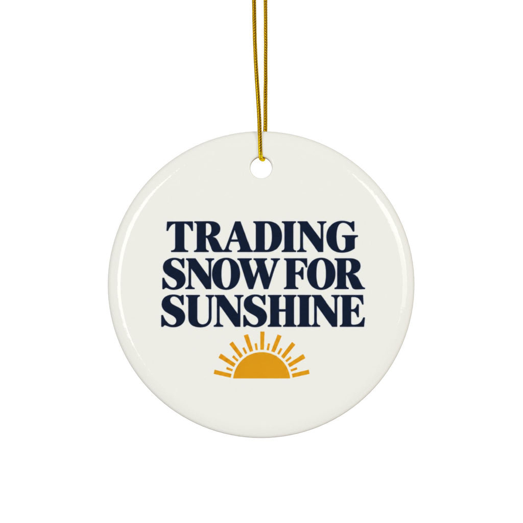 A round white ceramic ornament with a gold hanging string, featuring the text 'TRADING SNOW FOR SUNSHINE' in dark navy blue and a yellow-orange sun graphic at the bottom.