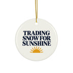 A round white ceramic ornament with a gold hanging string, featuring the text 'TRADING SNOW FOR SUNSHINE' in dark navy blue and a yellow-orange sun graphic at the bottom.