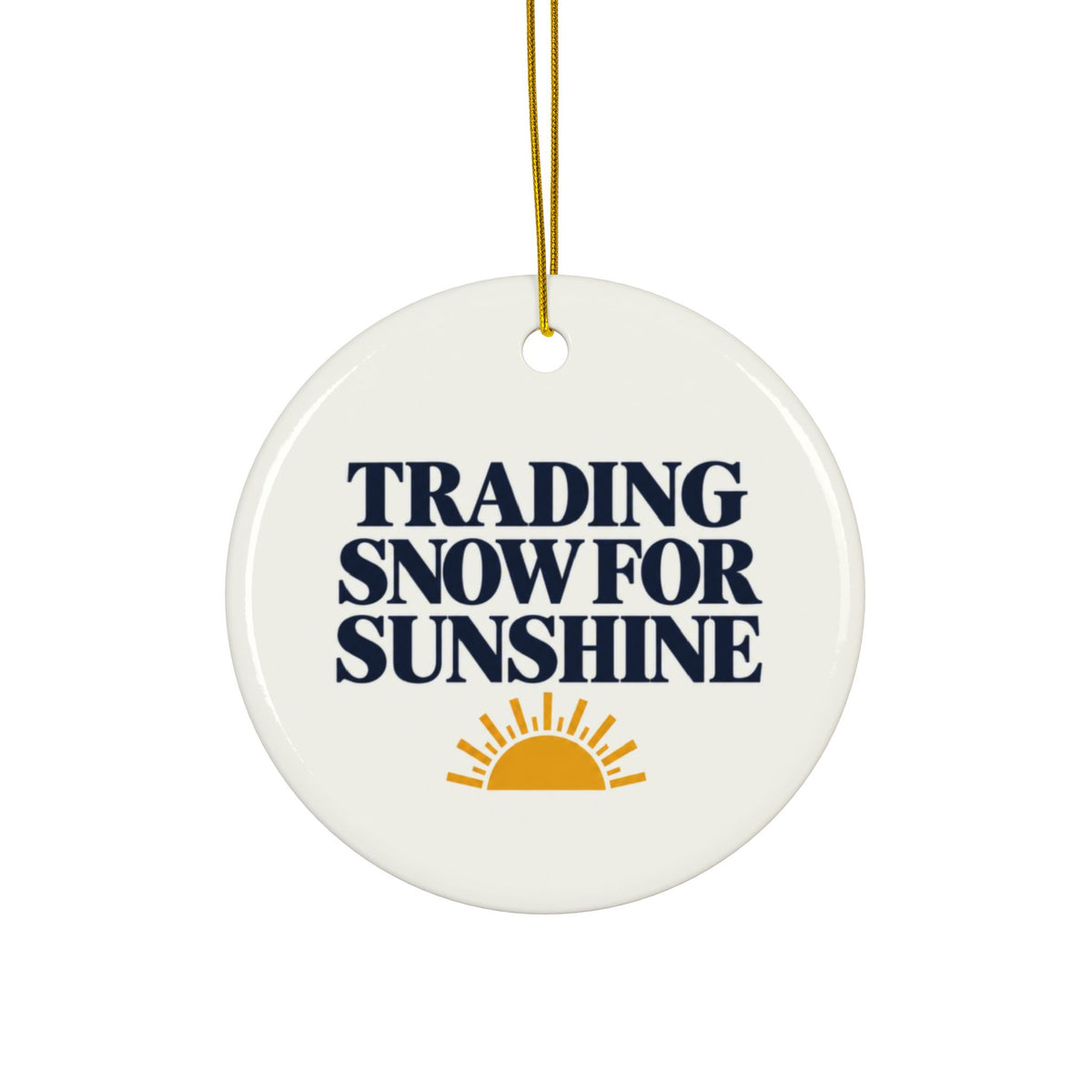 A round white ceramic ornament with a gold hanging string, featuring the text 'TRADING SNOW FOR SUNSHINE' in dark navy blue and a yellow-orange sun graphic at the bottom.