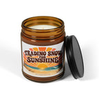 An amber glass jar candle with a black screw-on lid leaning against it on a white background. The front label reads 'Trading Snow For Sunshine' with a retro sunset and beach illustration, and small text below reads 'The Riverstone Life'.