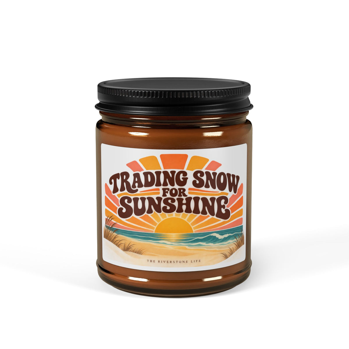 An amber glass jar candle with a black metal lid and a retro-style label reading 'Trading Snow for Sunshine' with a sunset and beach illustration with 'The Riverstone Life' words on the bottom. The candle is photographed on a clean white background.