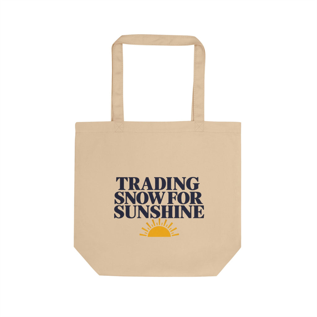 A beige canvas tote bag with two top handles, featuring the printed text 'TRADING SNOW FOR SUNSHINE' in dark navy blue and a small yellow sun graphic below, shown on a plain white background.