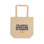 A beige canvas tote bag with two top handles, featuring the printed text 'TRADING SNOW FOR SUNSHINE' in dark navy blue and a small yellow sun graphic below, shown on a plain white background.