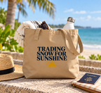 A beige canvas tote bag with the text 'TRADING SNOW FOR SUNSHINE' and a yellow sun graphic, styled on a beach with a straw hat, sunglasses, a water bottle, and a passport nearby.