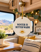 A round white ceramic winter ornament hanging from a wooden cabinet handle inside an RV or camper. The ornament features the text 'WINTER IS BETTER WARM' in navy blue lettering above a gold sun and waves graphic.