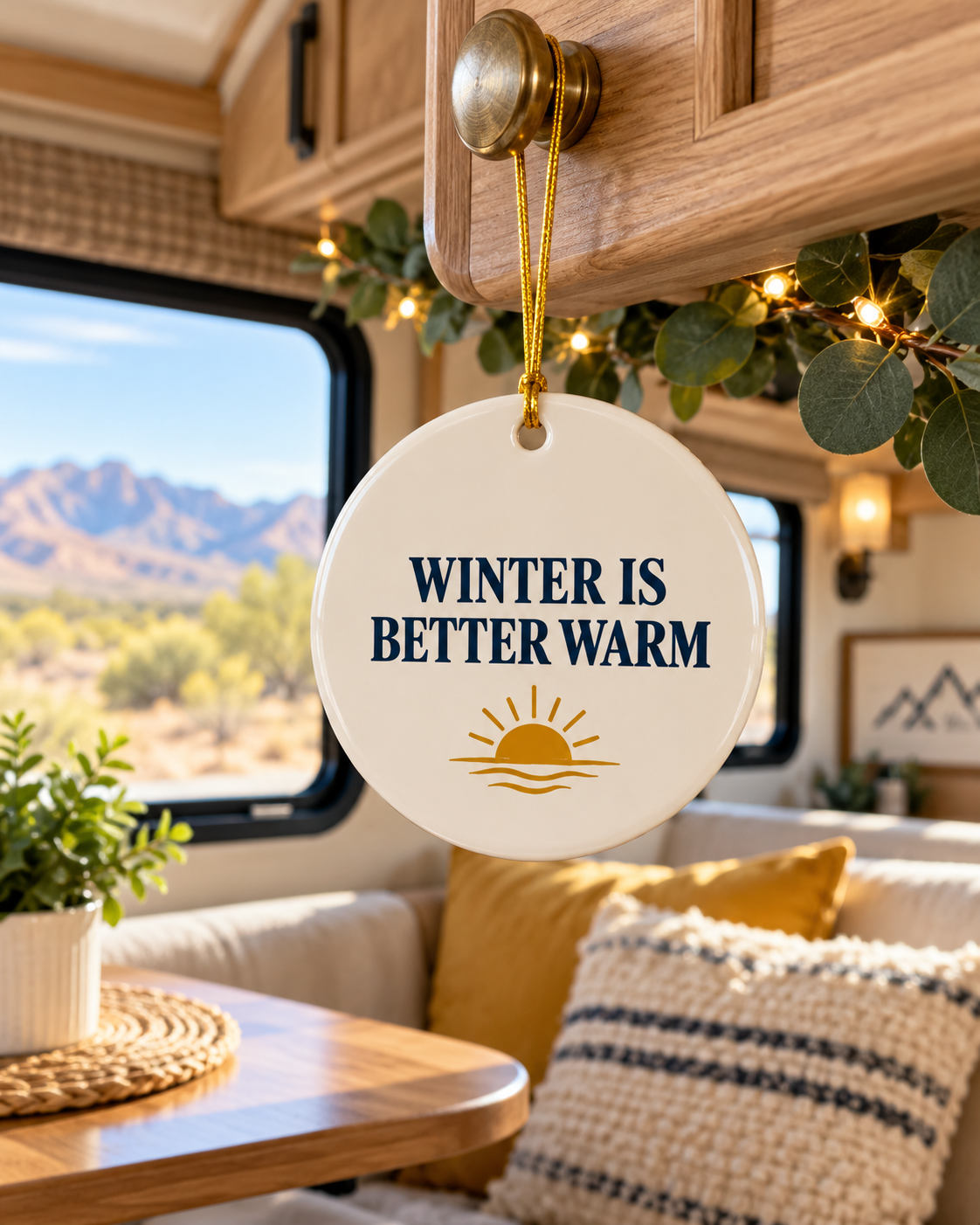 A round white ceramic winter ornament hanging from a wooden cabinet handle inside an RV or camper. The ornament features the text 'WINTER IS BETTER WARM' in navy blue lettering above a gold sun and waves graphic.