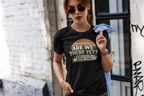 A woman wearing a black graphic t-shirt with a retro-style sunset design and the text 'ARE WE THERE YET? 47TH TIME', standing outdoors against a white brick wall.